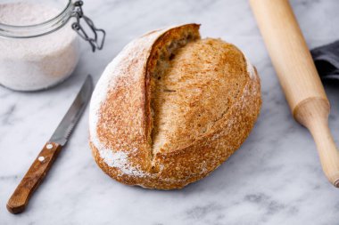 Çağdaş mermer masanın üzerinde un, bıçak ve ahşap raptiyeli bir somun ekşi ekmek. Mermer yüzeyde kırsal ekmek.  