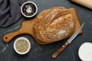 Kesme tahtasında bıçak olan taze pişmiş ekşi ekmeğin yuvarlak somunu, üst manzara. Siyah mermer masanın üzerinde tohumlu zanaatkar ekmeği. Rustik ekşi ekmek..