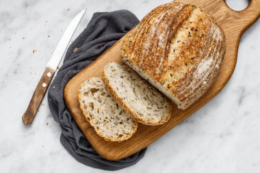 Yuvarlak somun taze pişmiş ekşi ekmek ve peçetede bıçak, üst manzara. Mermer masanın üzerinde tohumlu zanaatkar ekmeği. Peçeteye ekşi ekmek..