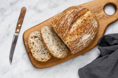 Kesme tahtasında bıçak olan taze pişmiş ekşi ekmeğin yuvarlak somunu, üst manzara. Mermer masanın üzerinde tohumlu zanaatkar ekmeği. Rustik ekşi ekmek..