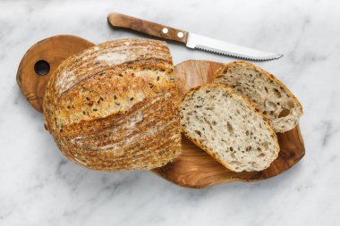 Kesme tahtasında bıçak olan taze pişmiş ekşi ekmeğin yuvarlak somunu, üst manzara. Mermer masanın üzerinde tohumlu zanaatkar ekmeği. Rustik ekşi ekmek..