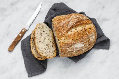 Yuvarlak somun taze pişmiş ekşi ekmek ve peçetede bıçak, üst manzara. Mermer masanın üzerinde tohumlu zanaatkar ekmeği. Peçeteye ekşi ekmek..