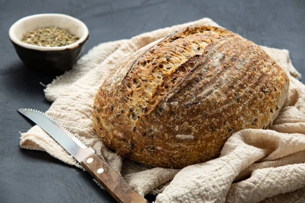 Round loaf of freshly baked sourdough bread with knife on napkin. Artisan bread with seeds on dark table. Rustic sourdough bread.