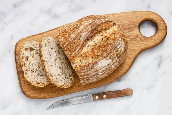 Round loaf of freshly baked sourdough bread with knife on cutting board, top view. Artisan bread with seeds on marble table. Rustic sourdough bread.