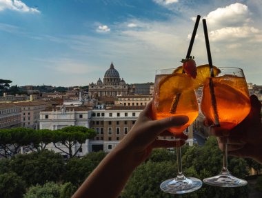 Travel photo concept. A couple is holding glasses of Aperol in the bar inside the Castle Sant'Angelo. Glasses of Aperol and St Peter's basilica at the background.  clipart