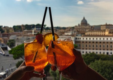 Beautiful travel picture in Rome. A couple holding glasses of Aperol Spritz. Cheering with glasses. City of Vatican is at the background clipart