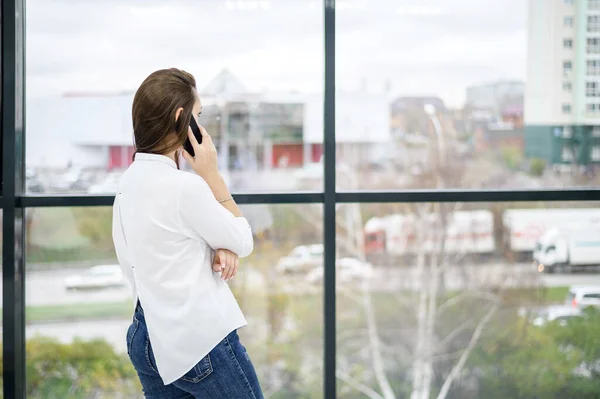 Una mujer con una camisa blanca está junto a la ventana y habla por teléfono. Chica europea está llamando a un teléfono inteligente y sonriendo . —  Fotos de Stock