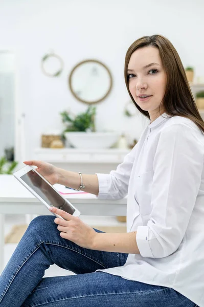 Una joven con una camisa blanca y jeans se sienta en una mesa con una tableta en las manos. Una mujer gerente de oficina realiza el trabajo en un dispositivo moderno . — Foto de Stock