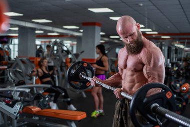 Spor salonunda sakallı kel bir adam. Halter egzersizi yapan kaslı vücutçu. Çıplak gövdeli güçlü bir adam. Genç sporcu halter yarışmalarına hazırlanıyor..