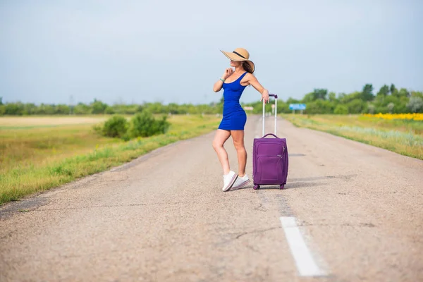 Μια γυναίκα στέκεται στο δρόμο με ένα καπέλο και μια μεγάλη τσάντα. Κορίτσι με μπλε εφαρμοστό φόρεμα με μωβ βαλίτσα στο δρόμο. Μια μελαχρινή με αθλητική φιγούρα περιμένει το λεωφορείο.. — Φωτογραφία Αρχείου