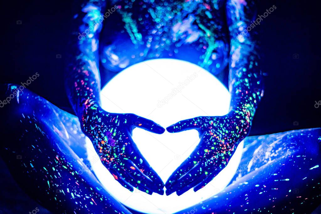 A woman painted with fluorescent powder holds a spherical lamp. Closeup of the legs of a girls hands with colored body spray. Round neon night light.