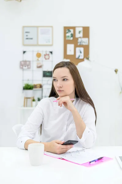 Una joven con una camisa blanca se sienta en un escritorio con un teléfono en las manos y planea un horario. Trabajadora de oficina hace un bolígrafo en el organizador. Cuaderno de papel para notas . — Foto de Stock