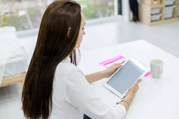 A young woman in a white shirt and jeans sits at a table with a tablet in her hands. A female office manager performs work on a modern device. The view from behind.