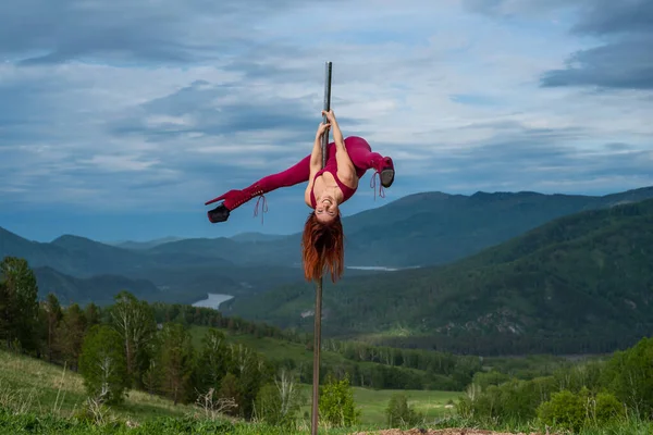 Güzel kızıl saçlı kadın dağların arka planında taşınabilir bir platformda dans ediyor. Kız direğin yanına cinsel olarak taşınıyor. Dersler için tulum. Yüksek topuklu. Altai. Müthiş esneklik. — Stok fotoğraf