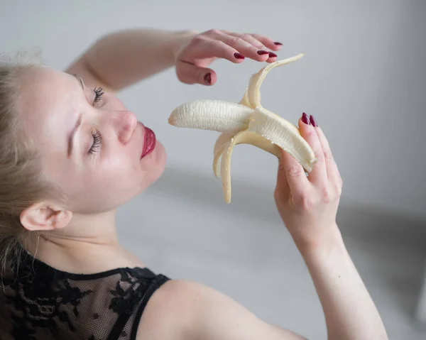 A loira em roupa interior de renda preta eroticamente come uma banana. Mulher atraente com lábios vermelhos sensuais suga sexualmente e lambe uma banana. Prazer oral. — Fotografia de Stock