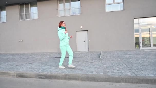 Uma mulher em um terno de esportes de hortelã quente e cachecol está andando pela rua e dançando. Menina ruiva feliz em uma camisola azul, calças e óculos caminha e abana espólio. Manhã gelada ensolarada cedo . — Vídeo de Stock