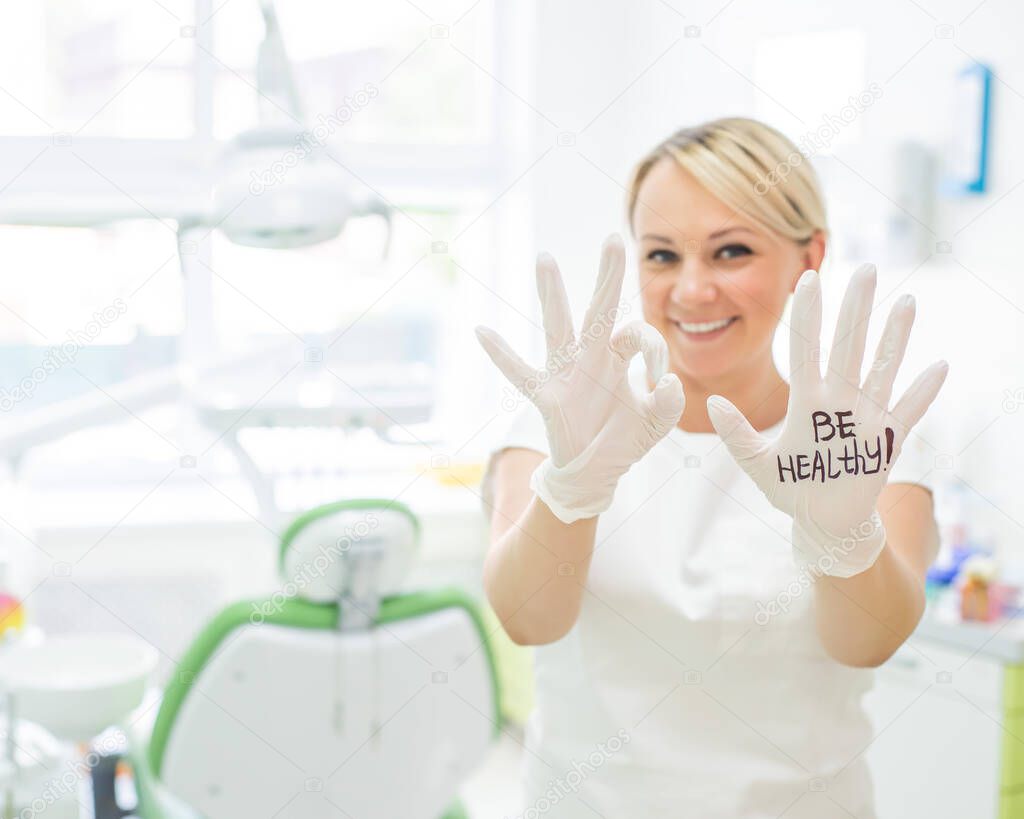 Friendly female dentist smiling and showing okay sign with fingers. Blonde doctor in gloves in the office in the medical center. The inscription on the hand be healthy