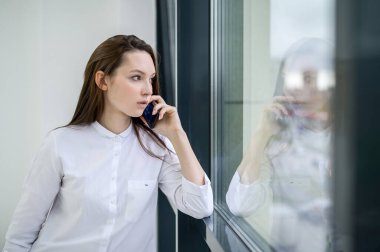Beyaz gömlekli bir kadın pencerenin yanında duruyor ve telefonda konuşuyor. Avrupalı bir kız akıllı telefondan arıyor ve gülümsüyor..