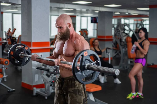 En skallig man med skägg i gymmet. Muskulös bodybuilder kille som gör skivstångsövningar. Stark man med en naken bål. Den unga idrottaren förbereder sig för tyngdlyftningstävlingar. — Stockfoto