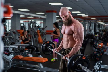 Spor salonunda sakallı kel bir adam. Halter egzersizi yapan kaslı vücutçu. Çıplak gövdeli güçlü bir adam. Genç sporcu halter yarışmalarına hazırlanıyor..