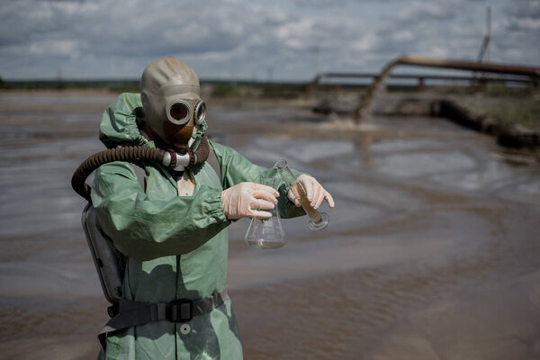 A scientist takes a sample of water from a reservoir after the release of chemical waste. A man in a respirator and a green protective suit from radiation. Water analysis experts. Mask.