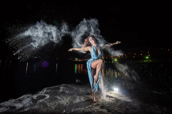 Femme rousse en robe bleue avec un décolleté profond dansant dans des nuages de farine. Une fille sur la rive de la rivière sautant éparpillant la poudre blanche . — Photo