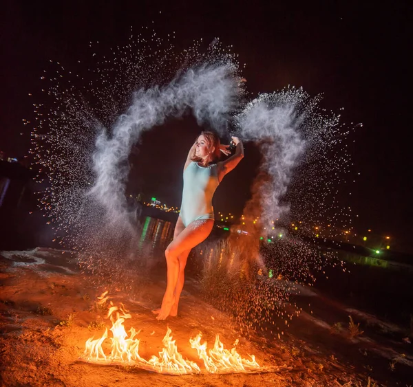 Femme rousse en maillot de bain bleu dansant dans des nuages de farine devant un feu. Fille sautant dispersant poudre blanche à côté d'une flamme brûlante . — Photo