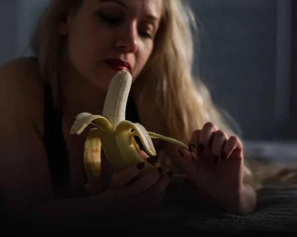 Die Blondine in schwarzer Spitzenunterwäsche isst erotisch eine Banane. Attraktive Frau mit sinnlichen roten Lippen saugt und leckt sexuell eine Banane. Mündliches Vergnügen. — Stockfoto