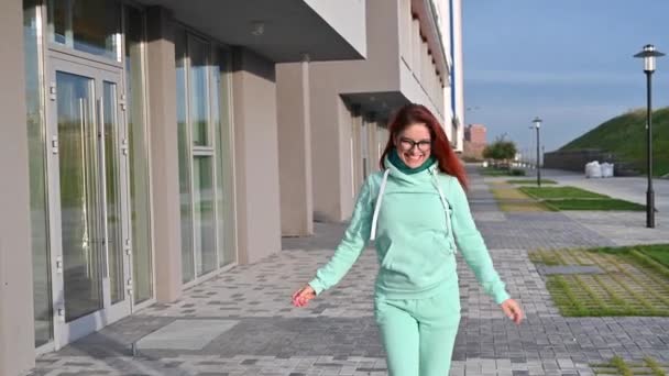 Una mujer en un cálido chándal de menta y bufanda camina por la calle y sonríe. Chica pelirroja feliz camina en una sudadera azul, pantalones y gafas. Temprano soleado mañana helada . — Vídeos de Stock