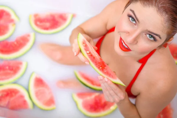 Porträt einer Frau im roten Badeanzug, die eine Wassermelone isst und grinst. Rotschopf Mädchen mit rotem Lippenstift nimmt ein Bad mit Milch und Früchten, um ihre Haut zu verjüngen. Körperpflege. — Stockfoto