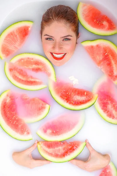 Portret van een mooie roodharige vrouw neemt een bad met melk en plakjes watermeloen. Spa behandeling voor huidverjonging en voeding. Rijpe plakjes van de grootste bes. — Stockfoto