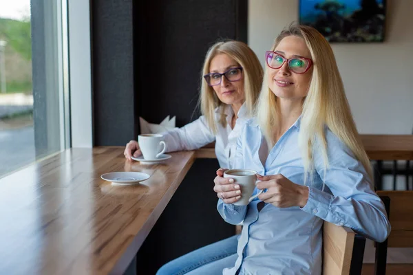 Смачна блондинка з окулярами і її старенька мати сидять у кафе і п'ють каву. День матері. Красива літня жінка та її доросла донька посміхаються та балакають. Схожість між ними. — стокове фото