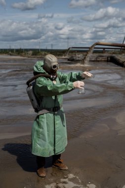 Yeşil koruyucu kıyafetli ve gaz maskeli bir erkek çevreci kirli bir gölde su örneği alır. Üretim israfı. Bir bilim adamı zehirli atık üzerine toksikolojik bir çalışma yapıyor..