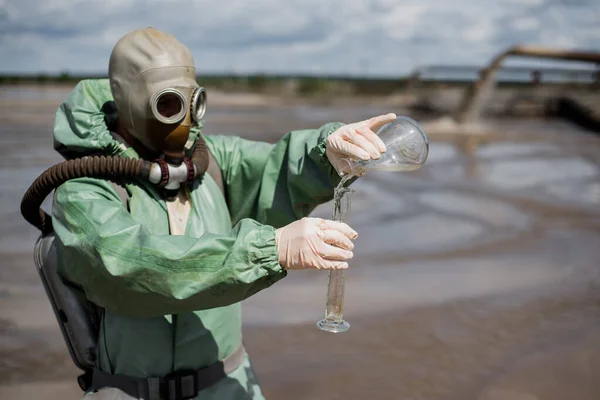 Mužský ekolog v zeleném ochranném obleku a plynové masce odebírá vzorek vody. Vědec právě provádí toxikologickou studii. Post apokalypsa. — Stock fotografie