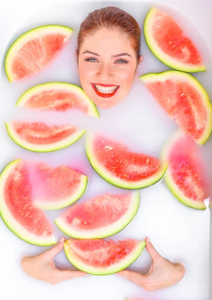 Portret van een mooie roodharige vrouw neemt een bad met melk en plakjes watermeloen. Spa behandeling voor huidverjonging en voeding. Rijpe plakjes van de grootste bes. — Stockfoto