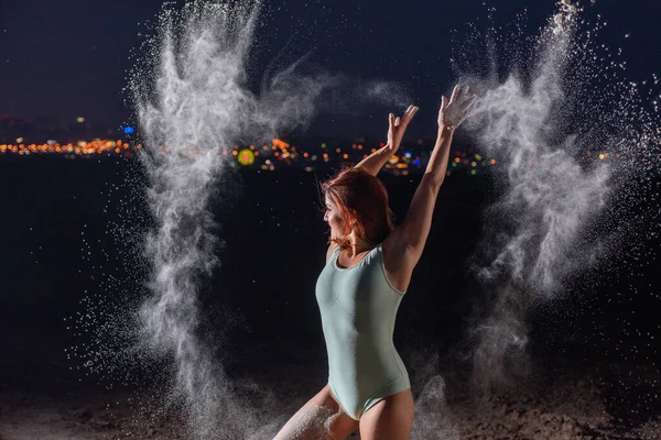 Rothaarige Frau im blauen Body, die in Mehlwolken tanzt. ein Mädchen am Flussufer springt und verstreut weißes Pulver. — Stockfoto