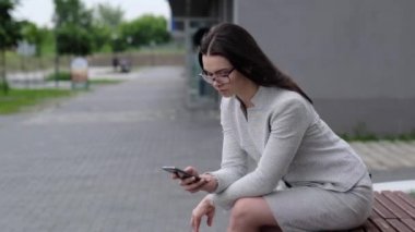 Ceketli ve etekli bir kız telefonda bir mesaj okuyor. Gözlüklü bir iş kadını, apartmanın avlusundaki bankta otururken akıllı telefonun ekranına parmağını sokuyor..