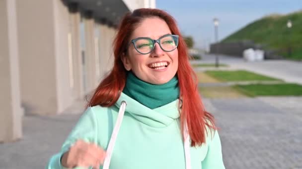 Retrato de una mujer en un acogedor y cálido chándal y bufanda camina por la calle y sonríe. Chica pelirroja feliz camina en una sudadera azul, pantalones y gafas. Temprano soleado mañana helada . — Vídeos de Stock