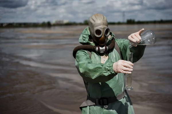 緑の保護服とガスマスクの男性環境保護者は、汚染された湖で水のサンプルを取ります。生産からの廃棄物毒性廃棄物の毒性研究をしています. — ストック写真