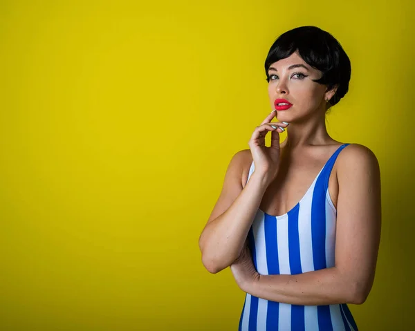Beautiful young woman in a black short wig and a striped swimsuit posing on a yellow background. Attractive girl with sensual lips is flirting at the camera. Bright makeup. — 스톡 사진