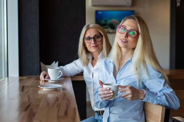 Смачна блондинка з окулярами і її старенька мати сидять у кафе і п'ють каву. День матері. Красива літня жінка та її доросла донька посміхаються та балакають. Схожість між ними. — стокове фото