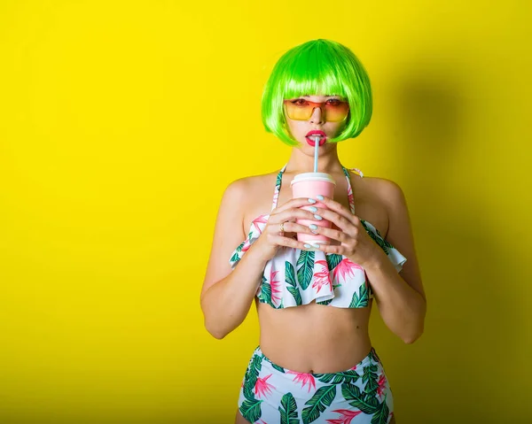 Beautiful young woman in a bright green wig under a quack and a swimsuit posing on a yellow background. Attractive girl with sensual lips flirts on the camera, drinks a drink through a straw. — 스톡 사진