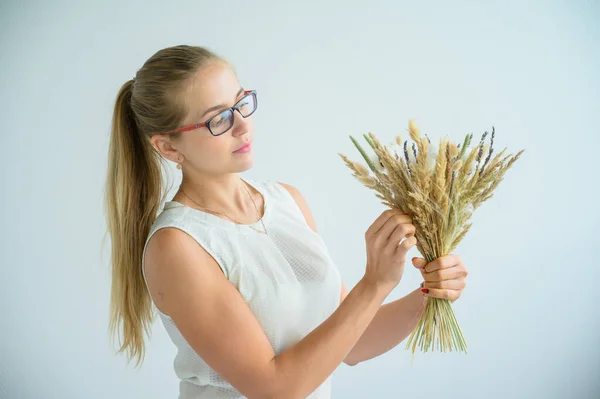 Die Blondine in Brille und weißer Bluse sammelt in einem weißen Atelier einen Strauß getrockneter Blumen und Baumwolle. eine charmante Floristin lächelt bei der Arbeit. — Stockfoto