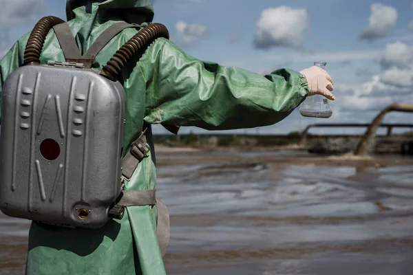 Mužský ekolog v zeleném ochranném obleku a plynové masce odebírá vzorek vody ve znečištěném jezeře pro výzkum. Produkce odpadu. Vědec provádějící toxikologické studie toxického odpadu. — Stock fotografie