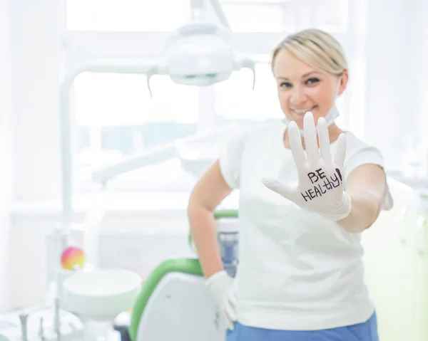 Amichevole dentista femminile sorridente e dando il cinque. Medico biondo con i guanti in un ufficio in un centro medico. L'iscrizione sulla mano essere sano . — Foto Stock