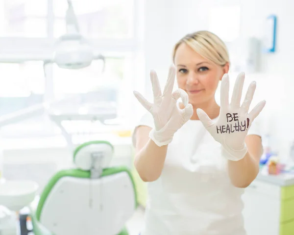 Odontóloga amigable sonriendo y mostrando buen signo con los dedos. Médico rubio en guantes en la oficina en el centro médico. La inscripción en la mano ser saludable —  Fotos de Stock