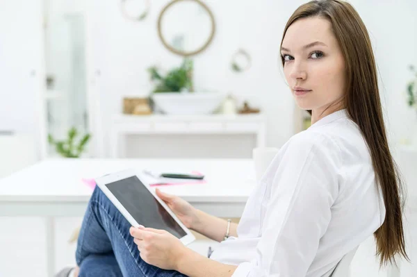 Eine junge Frau in weißem Hemd und Jeans sitzt mit einem Tablet in der Hand an einem Tisch. eine Bürokauffrau führt Arbeiten an einem modernen Gerät aus. — Stockfoto