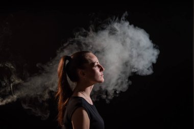 Esmer, siyah arka planda elektronik bir sigara içiyor ve ağzından yoğun bir duman salıyor. Bir kadın portresi sigara içiyor, havada süzülüyor..