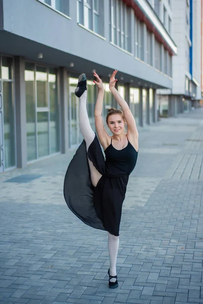 Bailarina en tutú posando contra un edificio residencial. Hermosa mujer joven en un vestido negro y zapatos puntiagudos saltando demuestra estiramiento. La bailarina está de pie en las divisiones. Afuera. . —  Fotos de Stock