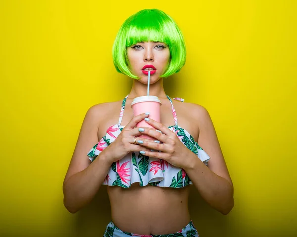 Beautiful young woman in a bright green wig under a quack and a swimsuit posing on a yellow background. Attractive girl with sensual lips flirts on the camera, drinks a drink through a straw. — 스톡 사진